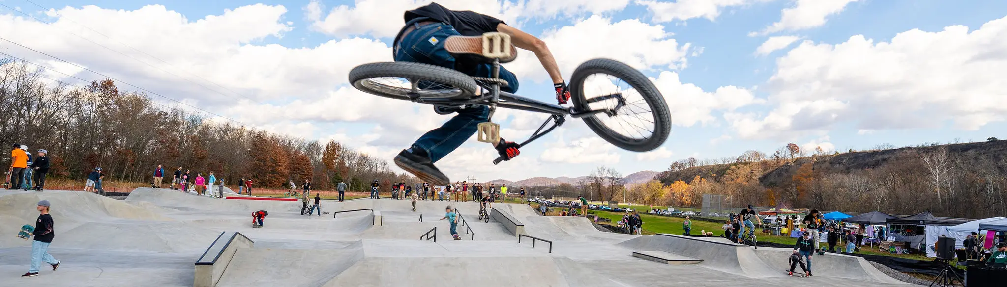 A BMX rider is in mid-air, performing a trick over a concrete ramp at a skate park with spectators watching from the sides